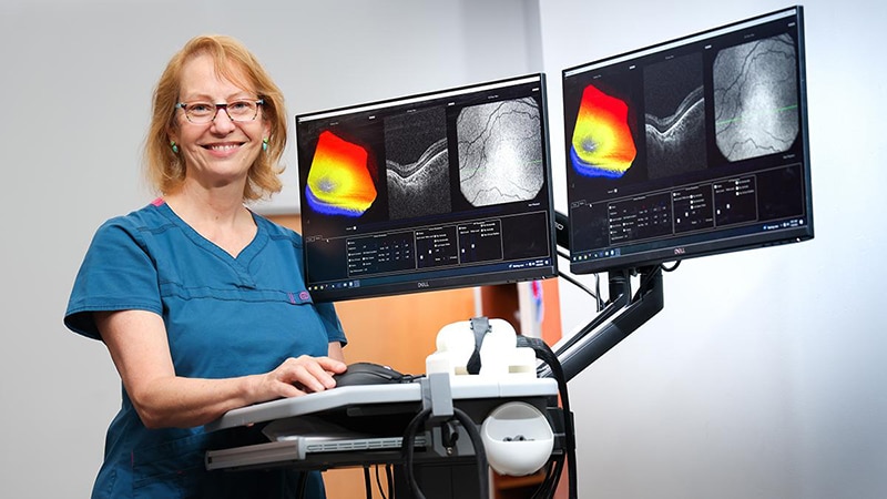 A woman in scrubs stands next to a device with two TV screens showing colorful images