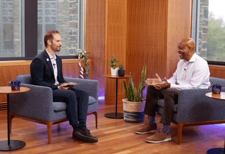 Rich Eva and Retired Gen. Charles Q. Brown Jr. sit across from each other in modern blue chairs during an interview in a well-lit room with wooden walls and large windows. They are smiling and engaged in discussion. A small table with plants and another with coffee mugs sit between them, and a Duke logo is visible on one of the mugs.